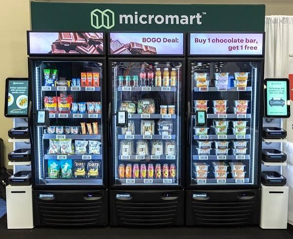 A single Micromart AI vending machine integrated into a stylish break area of a Kansas City tech startup office. The machine’s brushed metal frame and softly glowing interface are set against a backdrop of light wood cabinetry and dark quartz counters, with stainless mini-fridges and simple geometric decor nearby. Large windows reveal an abstract hint of Kansas City high-rises in the distance. Afternoon natural light washes in, supplemented by warm pendant lighting above, creating gentle highlights on the machine’s edges and a calm, productive atmosphere. Shot from a three-quarter angle with moderate depth of field, the photographic image feels clean, professional, and welcoming, illustrating convenient, smart vending as part of a modern workplace.