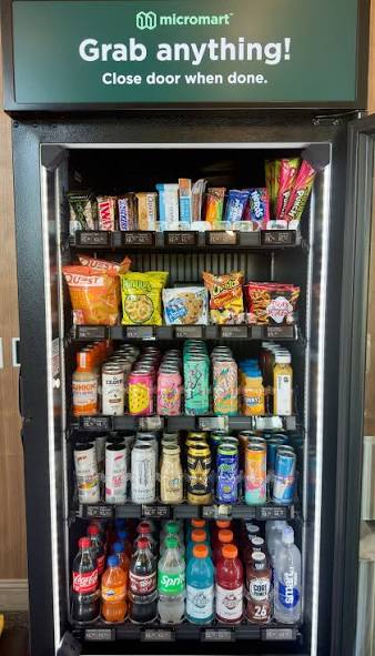 A sleek Micromart AI vending machine with a glossy black and brushed stainless steel exterior stands prominently in a modern corporate lobby in Kansas City, Missouri. The machine’s large, crystal-clear glass front reveals neatly organized snacks and chilled beverages in vivid packaging, with subtle digital interface elements glowing along the side. The polished tile floor reflects its clean lines, while blurred office doors and a city skyline through tall windows create context in the background. Soft, bright overhead lighting and natural daylight combine to create crisp reflections and gentle shadows. Shot at eye level with photographic realism, wide enough to show the environment but with the machine as the central focus, conveying professional, reliable, modern vending made simple.